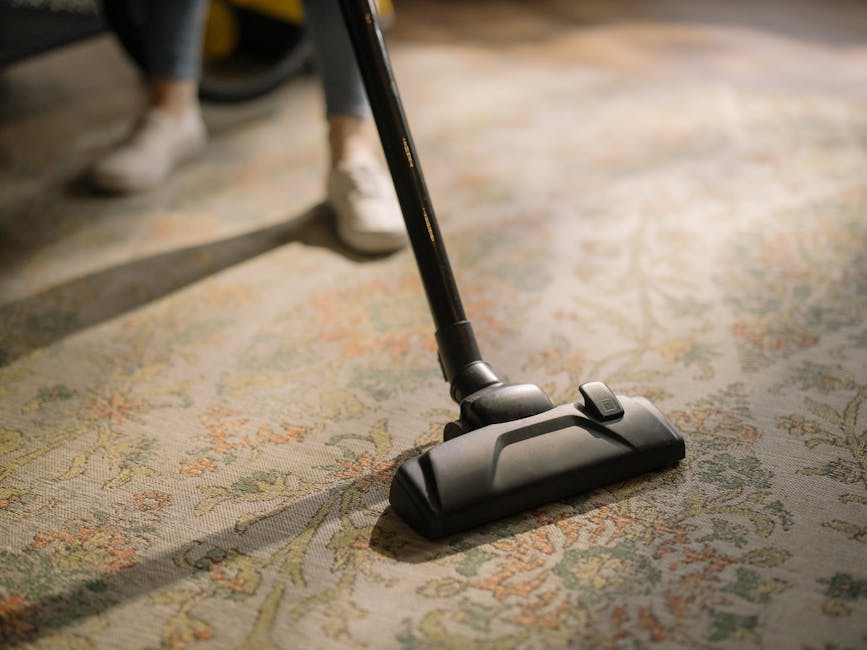 Close-up image of a black vacuum cleaner head with a handle, actively cleaning a patterned, cream-colored carpet in a room. The carpet features intricate floral designs in pastel green, orange, and beige tones. In the background, the lower legs and white shoes of a person are visible, indicating a domestic setting. Bright, natural lighting illuminates the scene, highlighting the cleanliness and condition of the carpet. The image underscores surface cleaning processes carried out by Carpet Cleaning Haringey, demonstrating professional deep cleaning for maintaining hygiene and surface appearance.