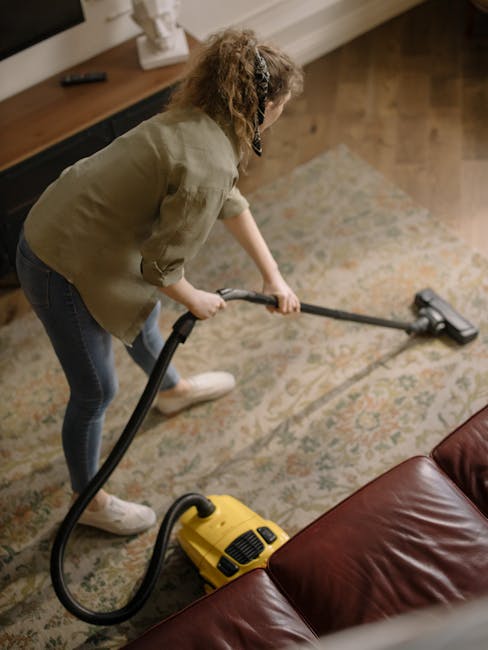 A woman engaged in domestic cleaning, using a vacuum cleaner to clean a patterned carpet in a living room. She is wearing a beige top and blue jeans, standing on a wooden floor next to a brown leather sofa. The carpet appears clean and well-maintained under the warm lighting, with a small side table visible in the background. The setting indicates a focus on surface cleaning and hygiene, typical of services provided by Carpet Cleaning Haringey, as highlighted in the Carpet cleaning Haringey Alexandra Palace results page. The vacuum cleaner's yellow body and black hose are prominent, and the room has a tidy, organized layout with a neutral color palette.