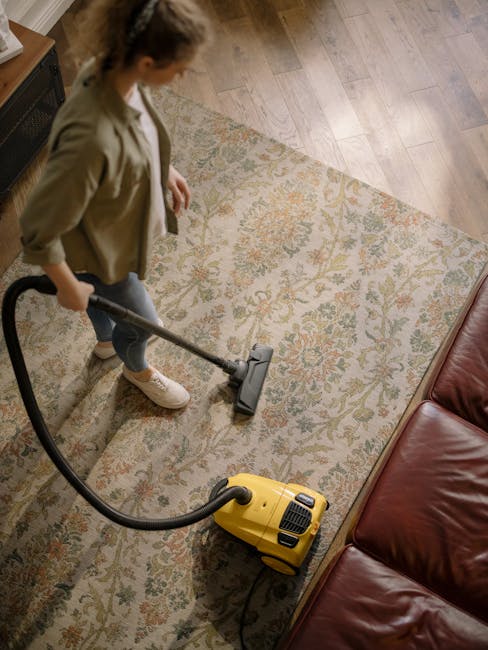 A person holding a yellow portable carpet cleaner with a black vent, preparing to operate it on a patterned, beige and green rug in a room with hardwood flooring and exposed brick wall. The scene emph