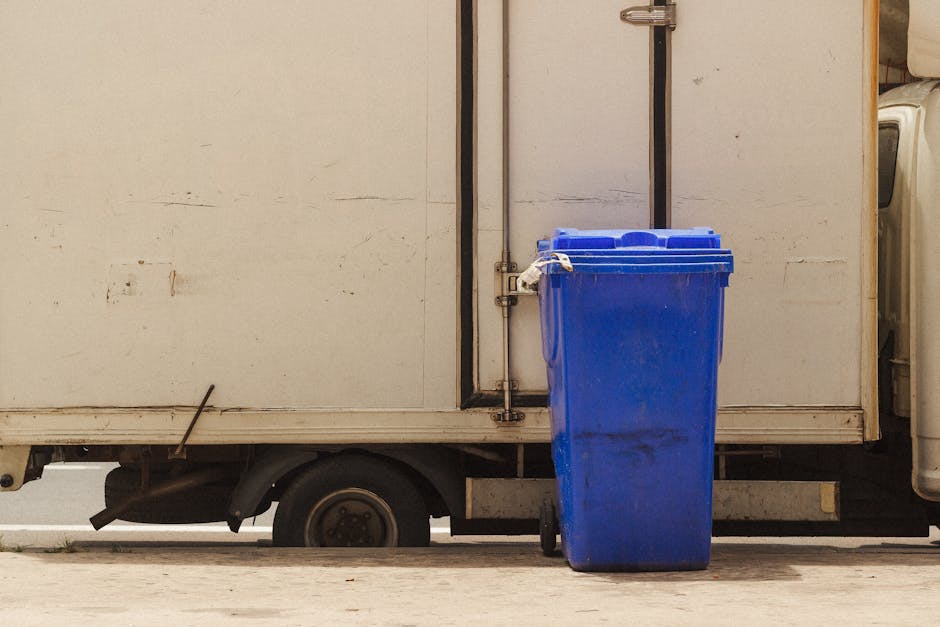 A person wearing a white long-sleeved shirt is disposing of crumpled paper waste into a round, wooden trash bin with a circular opening at the top, situated against a plain white wall. The environment
