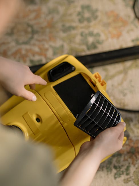 A person dressed in orange work overalls operating a yellow industrial vacuum cleaner with a large gray hose attached. The vacuum is positioned on a light-colored tiled floor inside a modern living ro