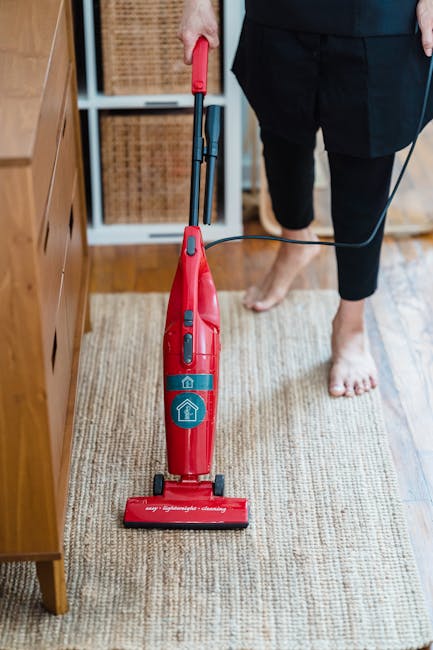 A woman using a yellow vacuum cleaner to perform surface cleaning on a patterned area rug in a living room. The rug features a floral design with muted colors, and the room has wooden flooring visible