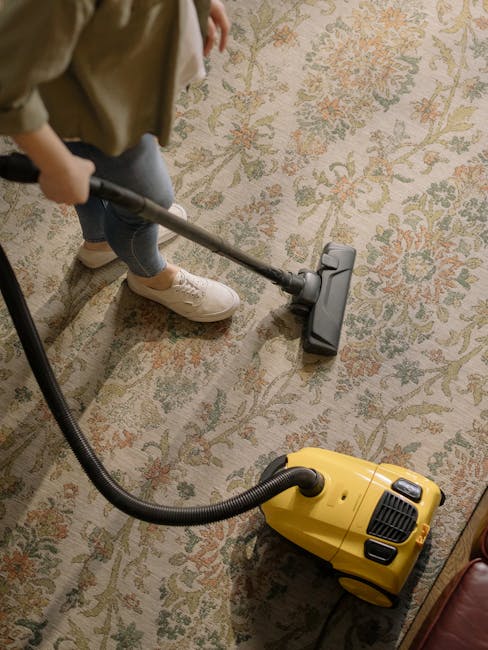A person, wearing beige trousers and white sneakers, is using a vacuum cleaner to deep clean a patterned carpet in a residential room. The vacuum cleaner, which is yellow with black accents, is placed on the light-colored, floral-printed surface. The individual is holding the extendable hose and cleaning the carpet, which appears to be in good condition with a soft texture. The room has ample lighting, with the carpet showcasing a design of floral motifs in muted tones of green, orange, and beige. This scene illustrates professional surface cleaning, focusing on hygiene and maintenance of domestic carpets. The image is associated with Carpet Cleaning Haringey, offering same-day emergency cleaning services in Tottenham.